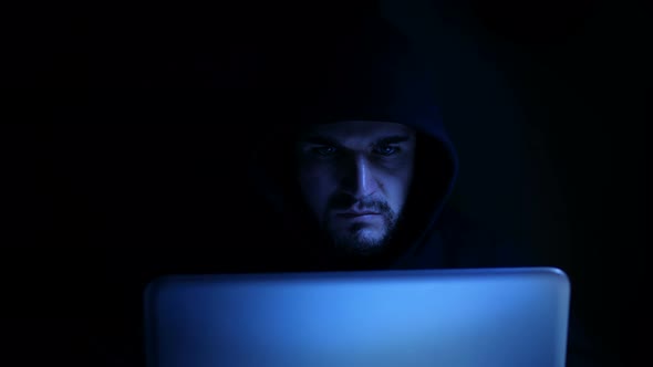 hacker with black hood types quickly on the computer at night