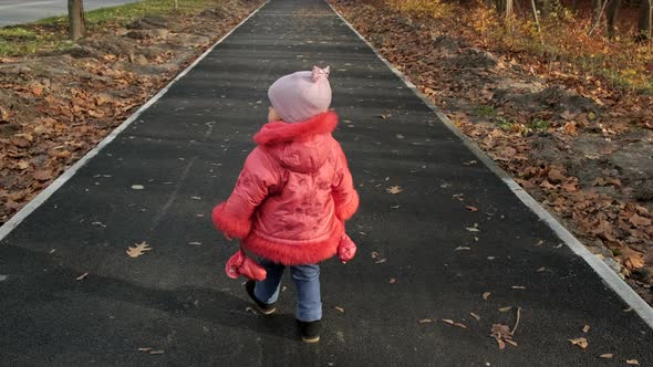 Little girl walks along the pavement along the park. View from the back, slow motion.