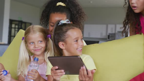 Video of diverse girls sitting at school common room with tablet, talking and laughing