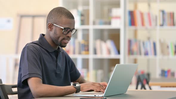 Young African Man with Laptop Smiling at Camera in Library