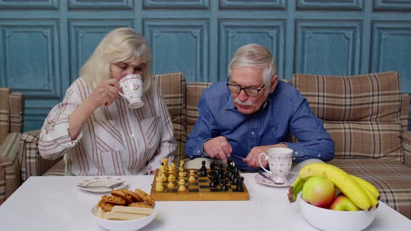 Smiling Senior Couple Grandfather Grandmother Resting on Sofa Drinking Coffee Playing Chess at Home