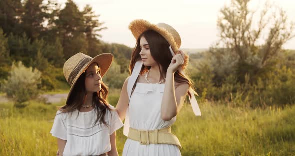 Two Girl Teenagers in White Dresses and Straw Hats with Flowers Bouquet Posing on Nature Landscape