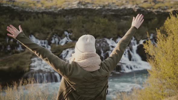 Back View of Young Traveling Woman Raising Hands Up and Enjoying the Beautiful View of Waterfalls in