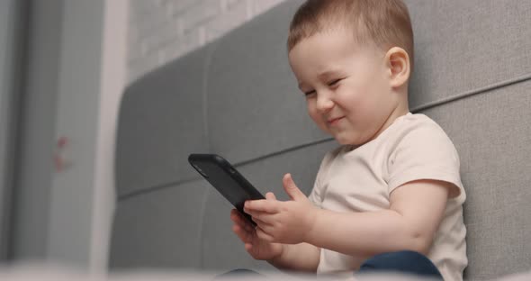 Little Boy Sits on the Bed Looking at the Smartphone and Smiling