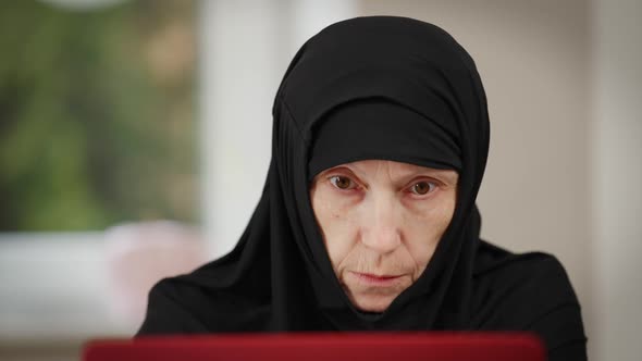 Front View Headshot of Concentrated Muslim Woman in Black Hijab Surfing Internet on Laptop Sitting