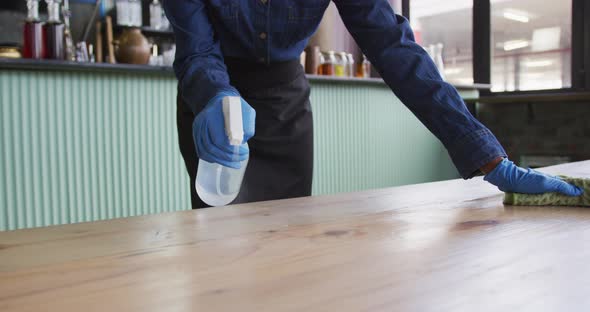 African american woman wearing gloves and apron disinfecting tables at cafe bar