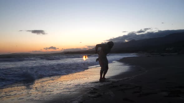 A man in silhouette dancing and spinning a burning fire staff on the beach at sunset with flames and