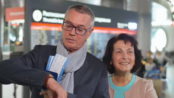 Elderly Couple Holding Tickets and Standing Over Information Board Background Waiting for Train