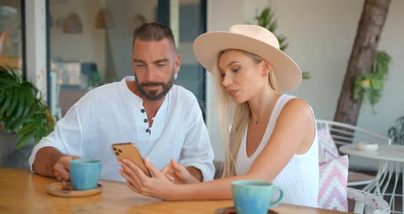 Young Woman in Hat Showing Photos on Her Phone to Young Man Sitting Outside in Spa Hotel Verandah