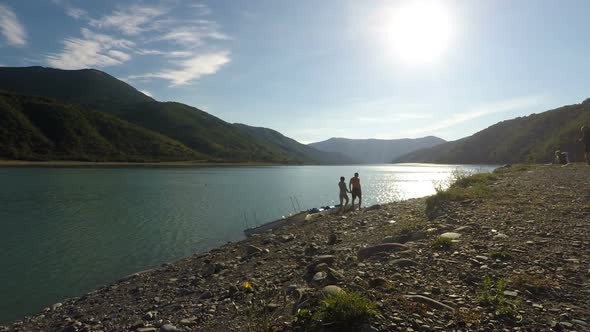 Couple Strolling Along Bank of Sunlit River Holding Hands, Mountains on Horizon