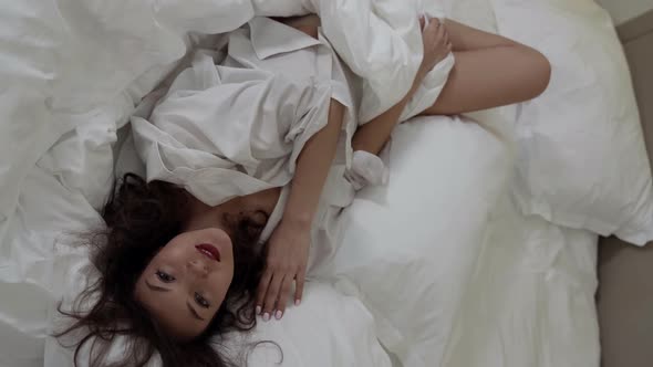 A Happy Brunette Woman in a Large White Shirt Playfully Poses in a Room on a Bed on White Bed Linen