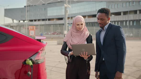 Female Dealer in Hijab Using Laptop While Showing Luxury Electric Car to Male