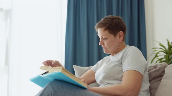 Relaxed Senior Woman Sitting on Sofa and Reading Book Indoors at Home