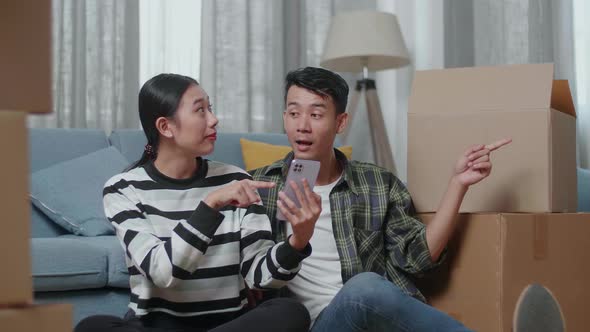 Young Asian Couple With Cardboard Boxes Look At Smartphone And Discuss About The New House