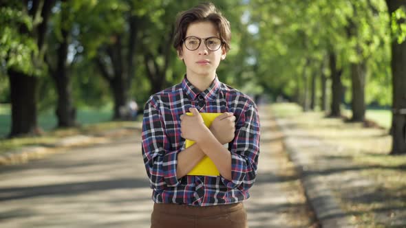 Portrait of Confident Smart Teenage Boy in Eyeglasses Posing in Sunny Summer Park with Book alt