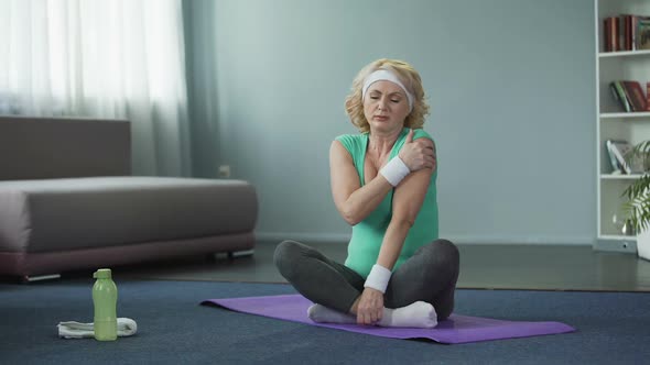 Blond Senior Woman in Sportswear Sitting on Yoga Mat and Massaging Her Shoulder