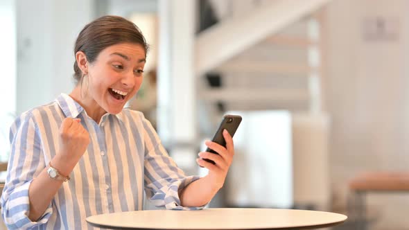 Excited Indian Woman Celebrating Success on Smartphone in Cafe 