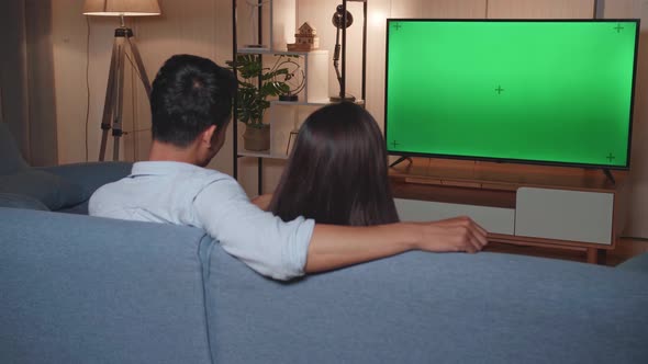 Back View Of Young Asian Couple Relaxing And Watching Tv With Mock Up Green Screen At Home