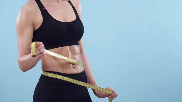 Woman an Athletic Body Measures Waist with a Meter Ribbon on Blue Background in the Studio