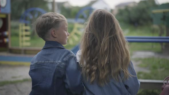 Positive Caucasian Cute Schoolboy Hugging Schoolgirl Sitting on Bench on Playground