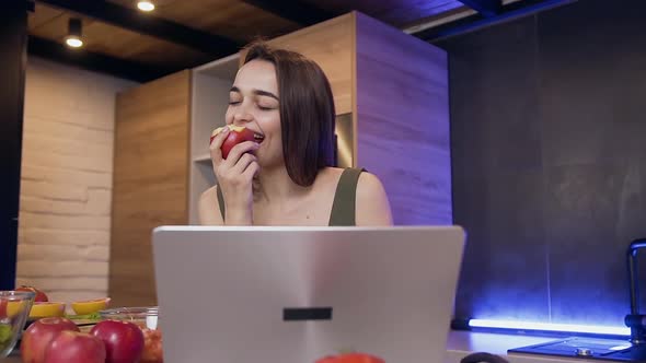 Smiling Girl which Eating Red Apple During Watching on the Computer in the Beautifully Kitchen