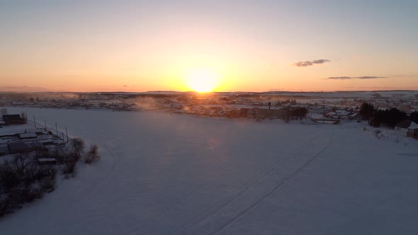 Aerial Over the Frozen Winding River Surrounded with Forest and with a Little Village on a Side