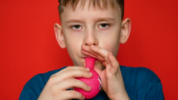 Portrait of a cheerful caucasian boy 7 years old inflating a balloon on a red background. studio sho