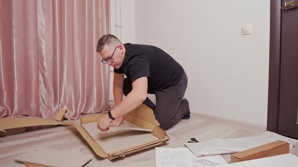 a Young Man Takes Out Parts for Assembling Furniture From a Box Assembles a Chest of Drawers on the