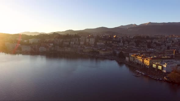 A drone view of a city surrounded by mountains, next to a lake, during a sunset in autumn. Lugano, T