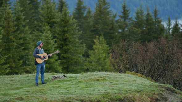 A man is playing the guitar at the top of a mountain surrounded by a forest