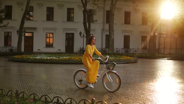 Young Woman Riding a City Bicycle with a Basket and Flowers in the City Center During the Dawn