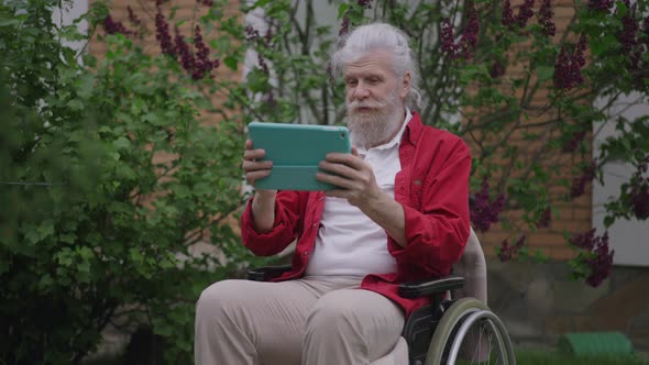 Positive Handicapped Greyhaired Caucasian Man Talking Using Video Chat on Tablet Sitting in