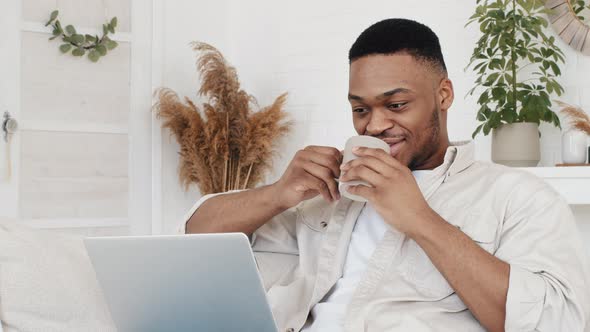 Young African Man Freelancer Afro Student Black Male Sitting at Home Couch Uses Laptop Computer