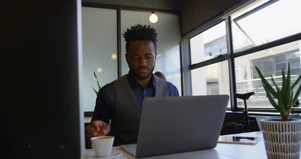 Front view of young black businessman working on laptop with a cup of coffee sitting at desk 4k