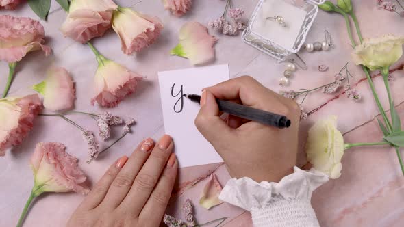 Hands writing YES I DO card near flowers close up on a marble table