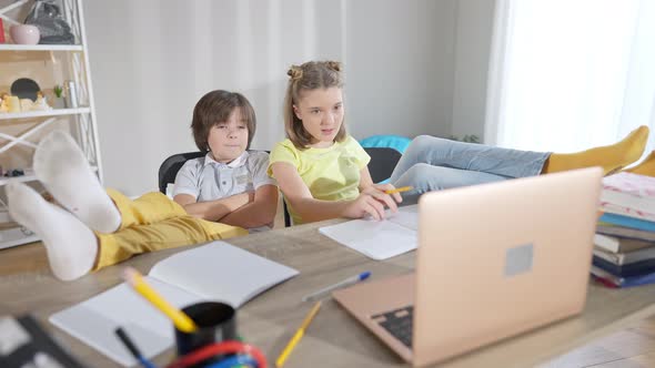 Wide Shot of Relaxed Caucasian Children with Feet on Table Looking at Laptop and Talking