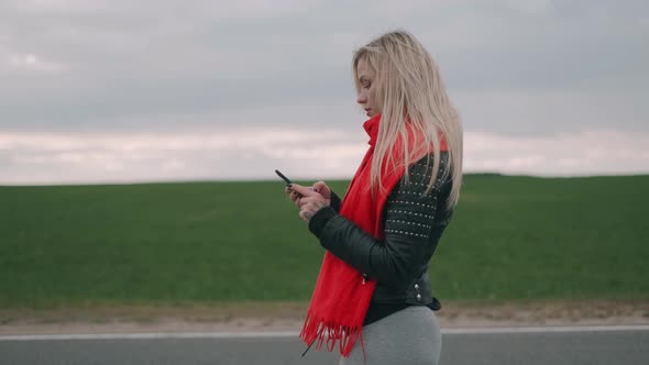 Blonde Woman Is Walking on the Road and Using the Smartphone. Girl with a Red Scarf Look at the