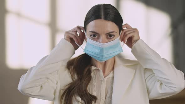 Portrait of Serious Young Millennial Businesswoman Looking at Camera Putting on Covid Face Mask in