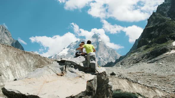 Married Couple of Tourists Sit on a Rock and Admire the Mountain View. Man Hugs a Woman in a Hike