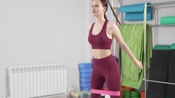 a Young Woman in a Tracksuit Does Squats with Gymnastic Elastic Bands