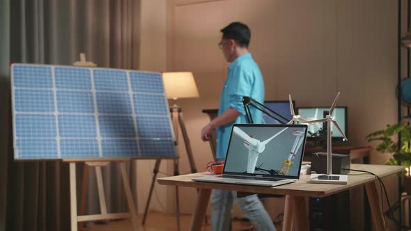 Asian Man Walks Into The Office That Has Solar Cell Next To The Laptop Showing Wind Turbine