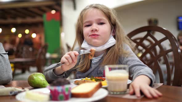 A Girl with a Protective Medical Mask on Chin Eats a Sausage in the Dining Room