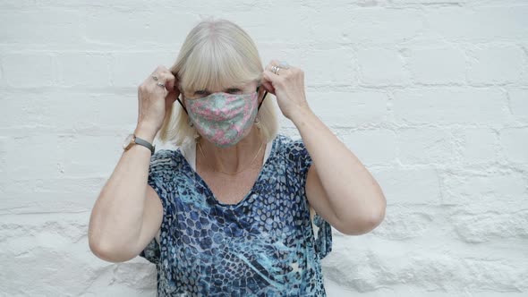 Elderly blond woman puts on floral face mask with a happy smile, safety concept for old people