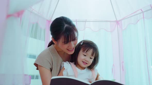 Asian mom read storytale, play education game to young baby girl daughter in tent at home.