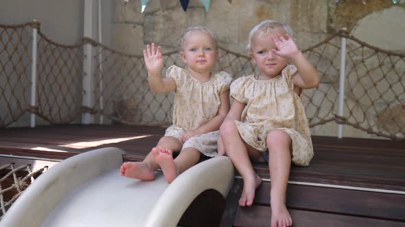 Twin Toddler Girls in Yellow Dresses Sitting on the Edge of the Slide and Waving to the Camera