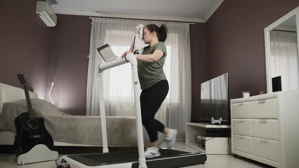 Chubby female is training at home, doing cardio fitness exercises on treadmill