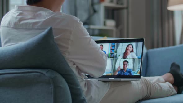 Man Greeting With Colleagues In Video Conference Call From Laptop While Lying On Sofa In The Living