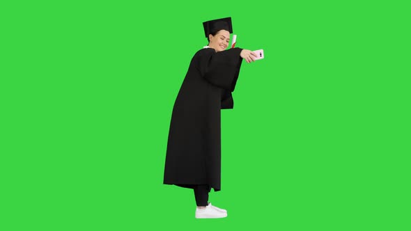 Happy Female Graduate Holding Diploma and Making Selfie on Her Phone on a Green Screen, Chroma Key.