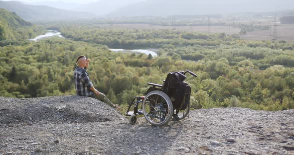Guy with Motionless Legs After Accident Sitting Near Wheelchair on the Top of the Hill