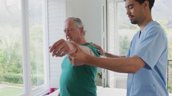 Biracial male physiotherapist slowing moving hand of senior man grimacing with pain at nursing home